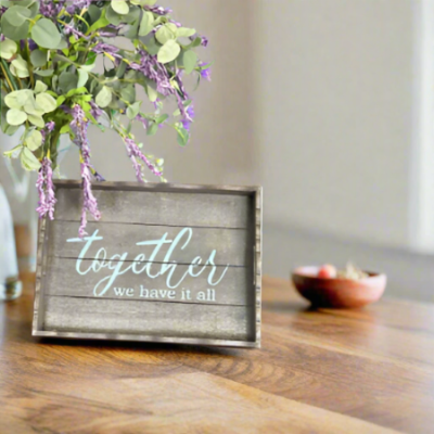 Decorative sign with 'together we have it all' text on a wooden surface with flowers in the background.